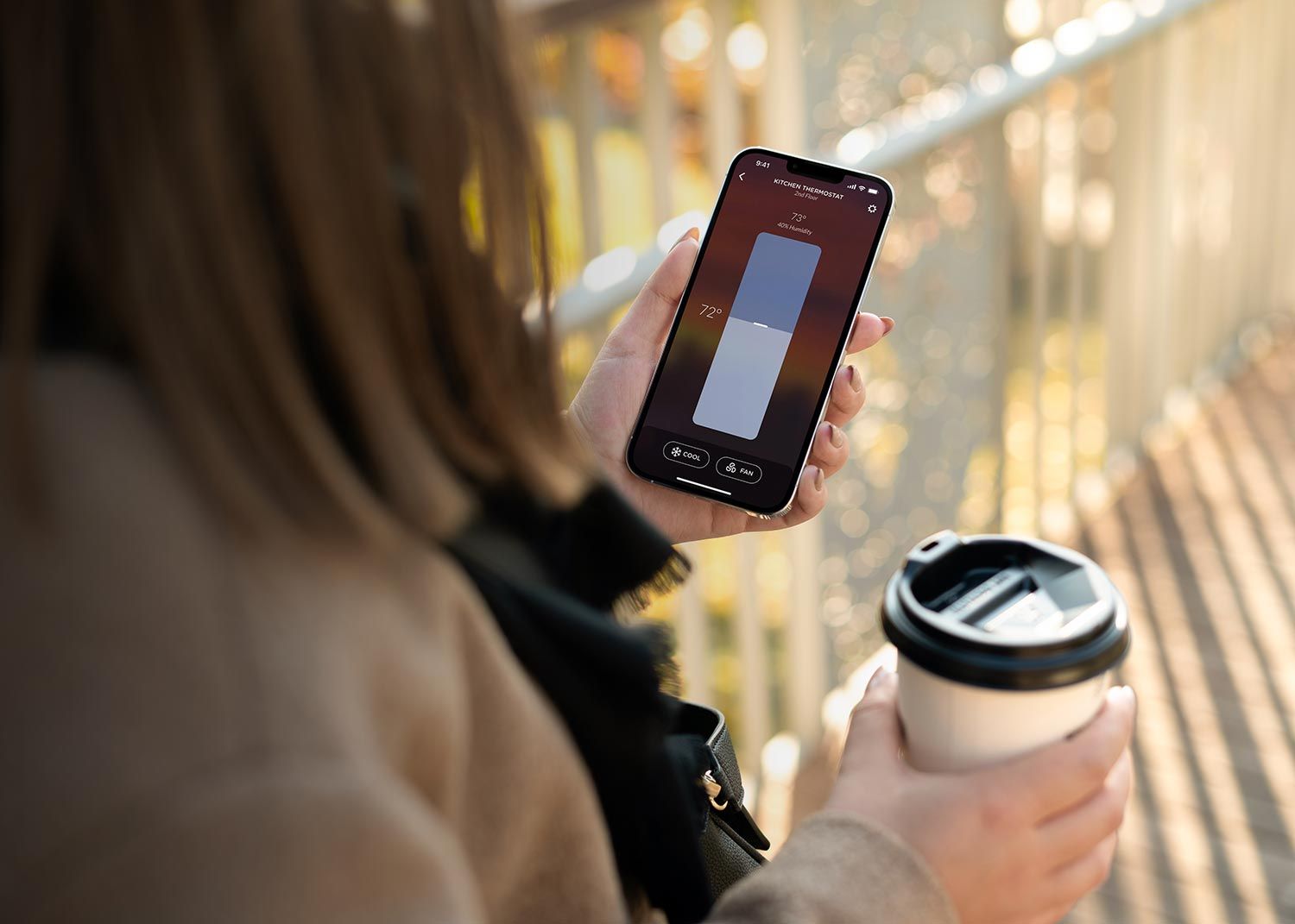 A person holding a smartphone with the Josh AI app open, showing thermostat controls, while holding a coffee cup, with a blurred outdoor background.